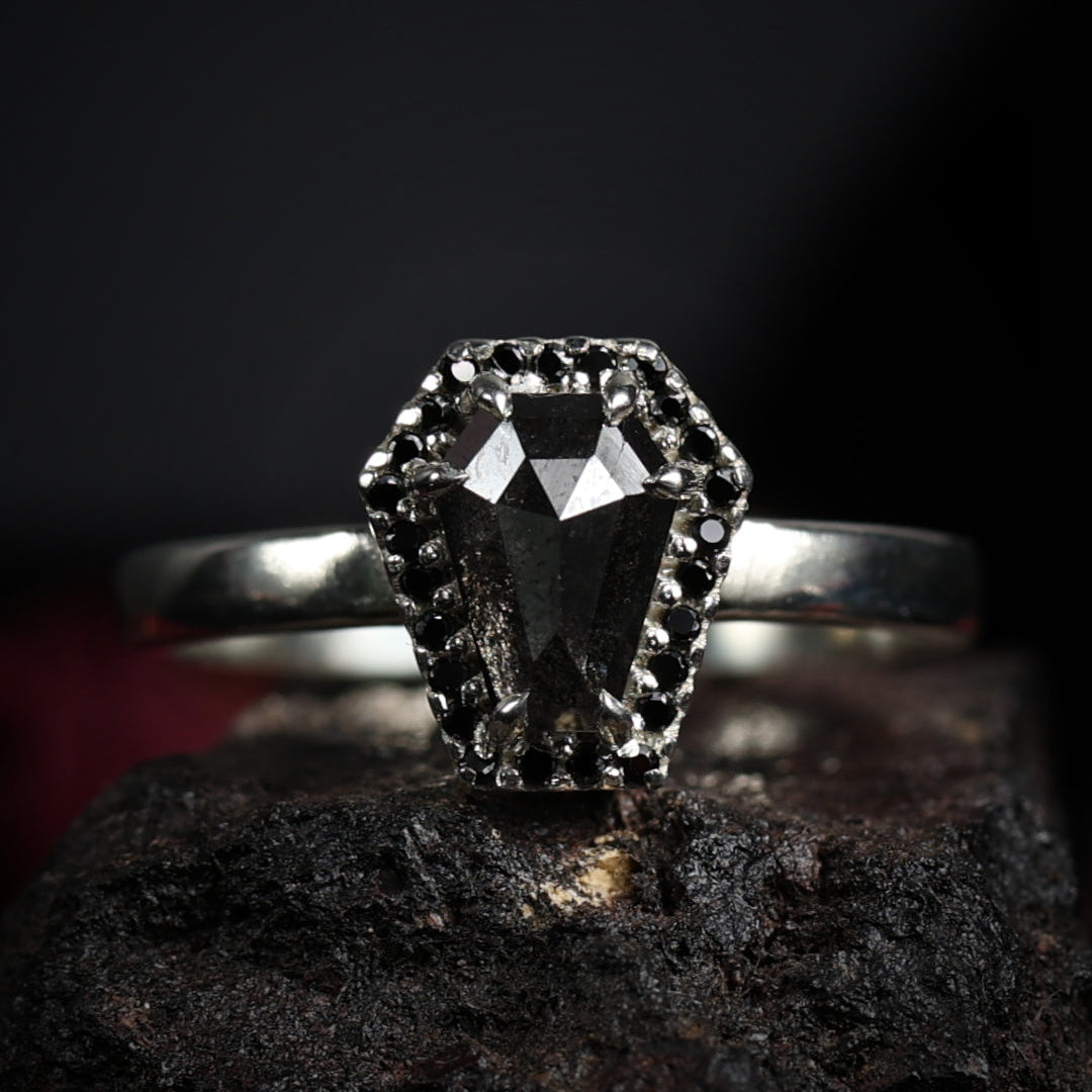 A gothic-style white gold ring featuring a large coffin-shaped salt & pepper diamond, surrounded by small pavé-set stones, displayed on a dark textured rock against a black background.