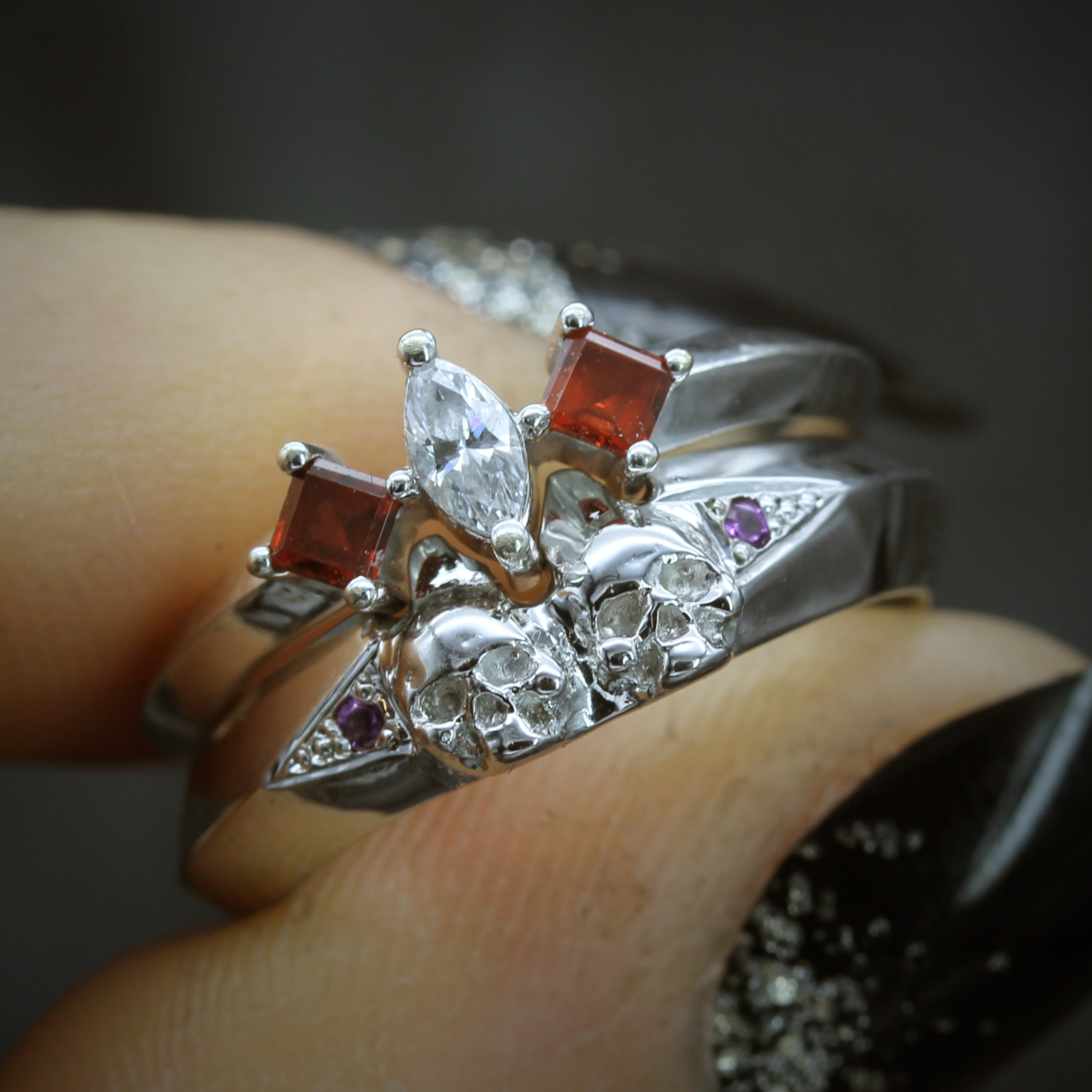 Paired ring stacked. Top is three stones ring, garnets shouldering diamond. Bottom ring is a double skull with amethyst shoulders. Held in finger.